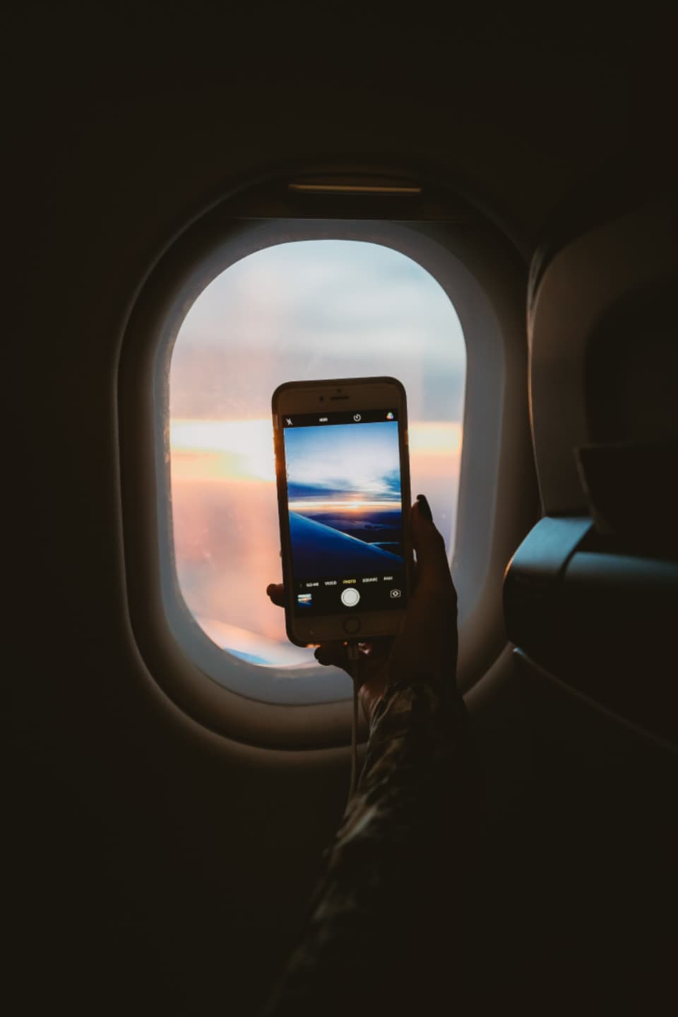 A hand holding a phone up to an airplane window at sunset, the phone's camera showing the clouds.